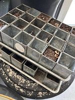 Metal seedling tray showing 24 square compartments with soil residue, placed on a rustic stool.