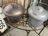 Photo showing two vintage ice buckets side by side on a metal table: the darker silver plated bucket with handles and the lighter hammered aluminum bucket.