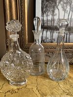 Photo showing all three vintage glass decanters with stoppers on a table in front of a framed picture.
