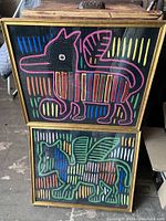 Full view of two framed vintage fabric art pieces stacked vertically showing colorful abstract animal figures on black fabric backgrounds with stitched details and wooden frames.