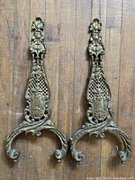 Pair of brass andiron end pieces shown lying flat on wooden floor showing front side and decorative details.