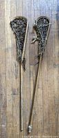 Two vintage wooden lacrosse sticks laid side by side on a wooden floor, showing front view of the frames and netting.
