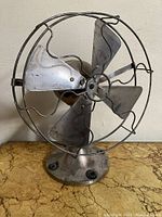 Front view of vintage metal fan showing four wide blades and circular protective wire cage.