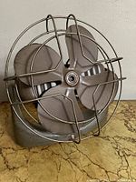 Front angle showing four-blade metal fan with round wire cage, placed on a flat surface.