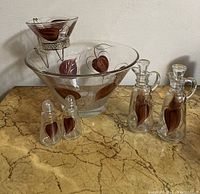 Entire set of vintage mid century modern glassware with brown leaf design including chip and dip bowl on metal stand, large serving bowl, salt and pepper shakers, and oil and vinegar bottles placed on marbled surface.