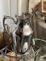 Full side view of the vintage chrome thermos carafe with curved handle and hinged lid, showing reflective finish and good condition.