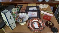 View of assorted Asian art items including porcelain vases, bowls, framed paper art, folding fan, carved stone figurines, and lacquer box arranged on table.