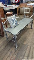 Metal bench with curved seat and open backrest, silver-gray finish shown from front angle with small decorative needlework pillow placed on seat
