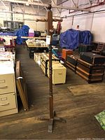 Full view of wood coat rack showing top hooks, central pole, and cross-shaped base in a warehouse setting.