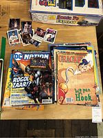 Top view of various vintage magazines and comics showing DC Nation comic and Cracked magazine with 'We Pan Hook' cover, spread out with sports cards on a table.