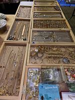 Ten wooden trays laid out on table filled with assorted costume necklaces, bracelets, rings and earrings
