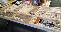 Wide shot showing multiple wooden trays with assorted costume jewelry including necklaces, earrings, rings, bracelets, pendants, and pins spread out on a table.