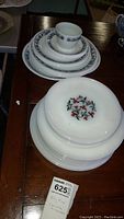 Stacked white Fire King plates showing red and green floral pattern in center, along with Corelle plates and cup featuring blue floral border.