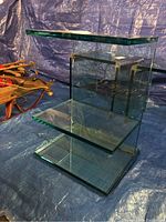 Glass side table with two shelves shown standing upright on a blue tarp background. Thick clear glass panels form top, bottom, and middle shelf surfaces.