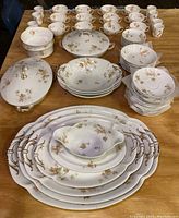 Full set on table showing multiple cups, saucers, plates, several covered serving bowls, and platters all with floral and leaf decorative pattern and gold accents.