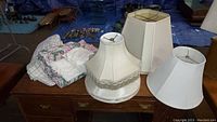 Five fabric lampshades displayed on a wooden desk with linens folded beside them