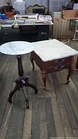Two marble top tables displayed on wooden floor: rectangular side table with ornate legs and drawer, and round pedestal plant stand with turned column base.