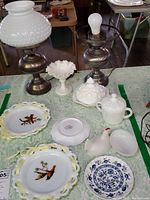 Overview of pair of electric lamps and milk glass dishes including candy/nut dishes, sugar bowl, hand-painted bird plates, and decorative items on a table