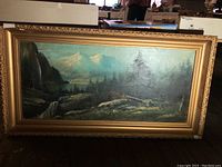 Full view of the landscape painting framed in a large gold decorative frame, showing mountainous scenery, a waterfall, trees, and possibly a lake in the background.