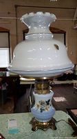 Full view of the lamp showing the ruffled white glass chimney and white milk glass shade with floral blue rose pattern, and part of the brass base.