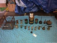 Wide view of 14 vintage oil cans with varied nozzles and 4 larger old stock motor oil cans on a blue quilted cloth.