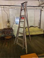 Full view of vintage orchard ladder in a room with antique furniture. Shows the A-frame wooden ladder with singular post stand and seven rungs.