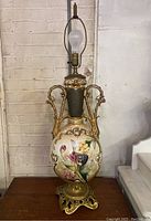 Full view of vintage ornate lamp with ceramic body and painted flowers, gold metal handles and base, angel molding at top.