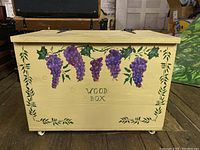 Front view of wooden box with grape painting and closed lid, showing the four caster wheels underneath.