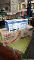 Photo showing the stacked boxes of Xerox fuser and Brother toner cartridge in an indoor setting with other items in background.
