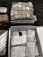 Assorted tile sheets and stacks including mosaic squares, rectangular tiles, and small brown textured tiles shown in a box and stacked on ground.