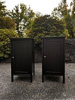 Front view of two matte black bedside tables side by side on pavement outdoors with shrubbery in the background.