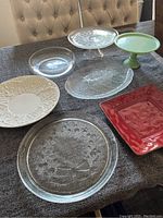 Seven assorted platters and cake stands arranged on a dark cloth-covered table: two cake stands (one green ceramic, one clear glass with holiday design), four distinct platters in cream, clear, and red colors.