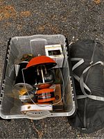 Plastic bin containing orange and black propane lantern with a glass chimney, various small camping accessories, and black carrying bag for tent.