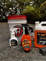 Photo showing Adam's Polishes wash bucket, Armor All Extreme Tire Shine in orange spray bottle, Meguiar's Gold Class Car Wash bottle, glass cleaner spray, and black sponge on ground.