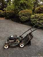 Side view of green Craftsman 6.0 HP push mower showing metal deck, wheels, handlebar, and attached grass catcher bag, placed on asphalt with bushes in background.