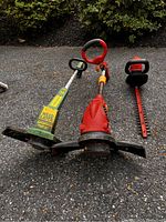 Front view of two electric weed eaters and one electric hedge trimmer laid on asphalt