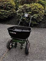 Front view of Scotts Elite fertilizer and seed spreader showing the black plastic hopper and metal frame with large wheels.