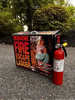 Image of the Kiddie Fire Escape Ladder box next to a small red fire extinguisher on pavement with trees and bushes in background.