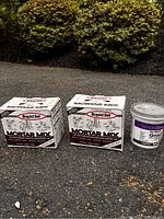 Two Rapid Set Mortar Mix boxes and one Simple Grout Pre-Mixed Grout bucket placed outdoors on a driveway with bushes in the background.