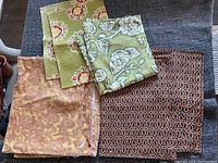 Three sets of assorted placemats and a folded green floral tablecloth laid out on a table.