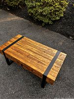 Full view of the rectangular rustic modern wood coffee table outdoors, showing wood surface and black metal legs and straps.