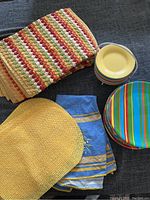 Set includes yellow woven placemats, multicolor striped placemats, yellow small plates stacked, multicolor striped dinner plates, and blue tablecloth with yellow border.