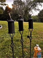 Photo showing three black metal tiki torches standing upright in grass with an unopened bottle of Tiki fuel next to them