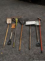 Photo shows seven assorted yard tools lined up on asphalt, including shovels, rakes, brooms, and a snow shovel, all showing wear and rust.