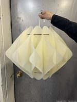 Full view of vintage off-white geometric plastic lamp shade being held by its cord against a metal door background.