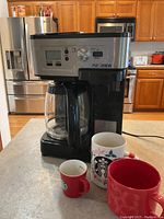 Hamilton Beach FlexBrew coffee maker on kitchen counter with glass carafe and three Starbucks coffee cups of varying sizes in front: small red espresso cup, white festive tall cup, and large red mug.