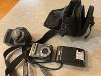Two Kodak Easy Share digital cameras and accessories on table including Z1285 with strap, DX6490, charger, battery, and camera case