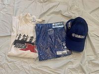 Photo displaying three clothing items: Yamaha licensed FX t-shirt still sealed in original blue plastic packaging, off-white used RacerX t-shirt laid flat, and navy blue Yamaha baseball cap with visible logos.