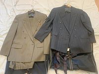 Two men's wool suits laid out, one olive green and one dark gray, showing jackets and pants with suspenders and pocket scarf on display.