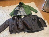 Three men's jackets laid out: green Columbia shell jacket opened to show fleece liner, black and gray NRA polyester shell jacket, brown Corporate Identity Apparel polyurethane coat with fur collar
